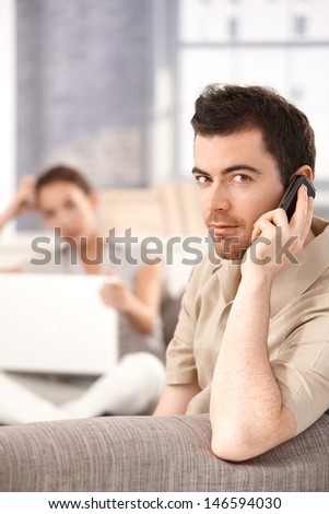 Young man sitting on sofa at home, talking on mobile, woman using laptop in the background.