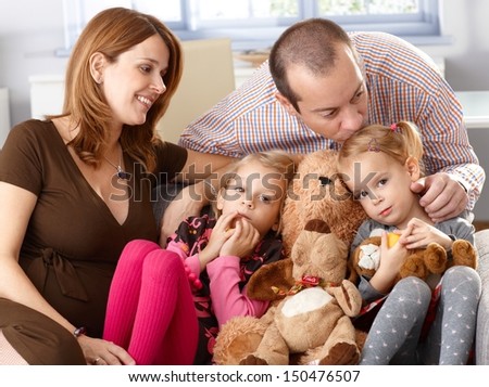 Family with two daughters and pregnant mother sitting on sofa at home, father kissing daughter.