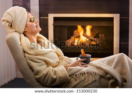 Young woman in cosy living room enjoying winter relaxation with face pack and tea, smiling.