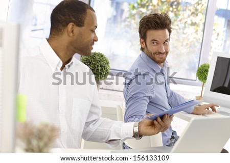 Young male office workers sitting at desk, one passing folder to the other in bright office.