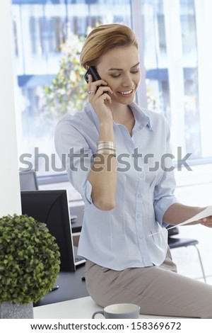 Happy young businesswoman talking on mobilephone, sitting on top of desk.