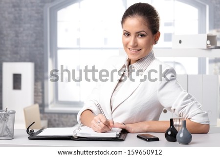 Portrait of young smart businesswoman writing notes, smiling at camera.