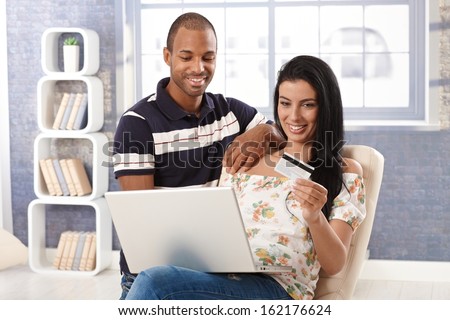 Diverse couple e-shopping at home, using credit card, smiling.