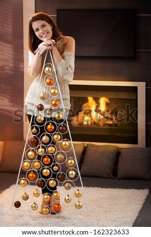 Happy attractive woman laughing, standing by design christmas tree at home, by cosy fireplace.