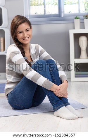 Full size portrait of beautiful young woman sitting on floor at home, arms folded around knee, smiling happily.