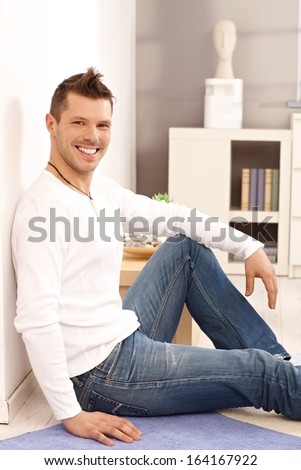 Happy young man sitting at home on floor, leaning to wall, smiling.