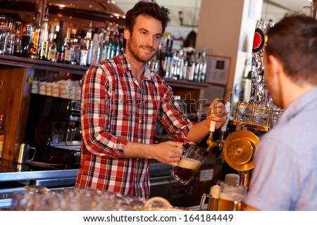 Portrait of young bartender serving beer in pub, looking at customer, smiling.