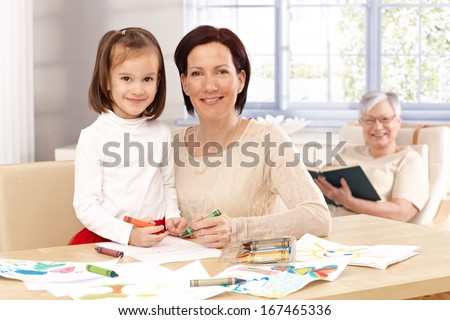 Happy mother and little daughter drawing at home, granny watching from background.
