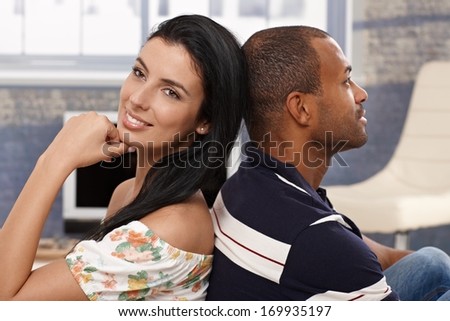 Happy interracial loving couple sitting at home back-to-back, smiling.