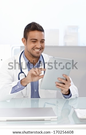 Happy male doctor using tablet computer, sitting at desk.