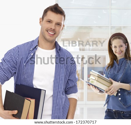 Male college student holding books at library, smiling, looking at camera.