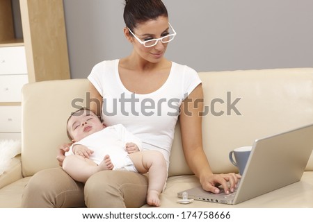 Young woman working on laptop computer while holding sleeping baby in arms.