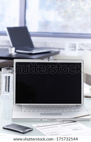 Closeup photo of open laptop with blank screen placed on desktop in bright office. Mobilephone and business report also visable.