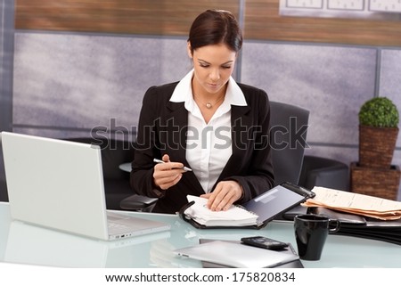 Young businesswoman looking at organizer, sitting at desk, working.