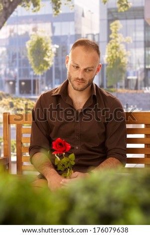 Disappointed man sitting on bench with red rose in hand.