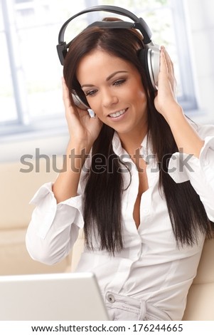 Young woman listening to music on headphones, using laptop computer, smiling.