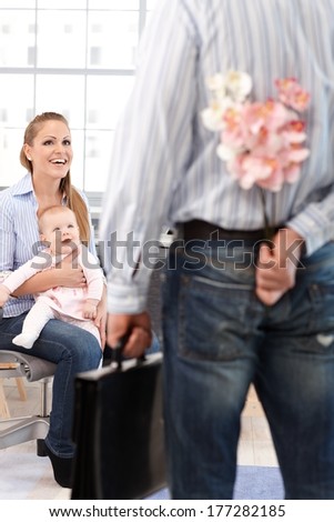 Woman sitting on chair with little daughter on lap, smiling happy at husband arriving home from work with flower hidden behind back.