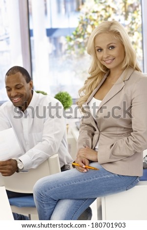 Attractive blonde office worker sitting on top of desk, smiling.