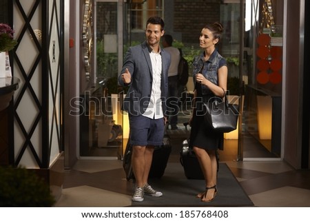Young couple arriving at hotel lobby, having suitcases, smiling, looking for reception desk.