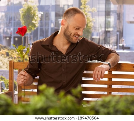 Young man waiting for date outdoors on a bench, holding red rose in hand, checking time on wristwatch.