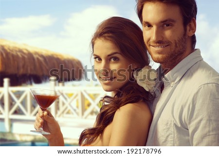 Young casual attractive caucasian lady with cocktail drink in hand and handsome boyfriend at tropical holiday beach. Smiling, looking at camera.