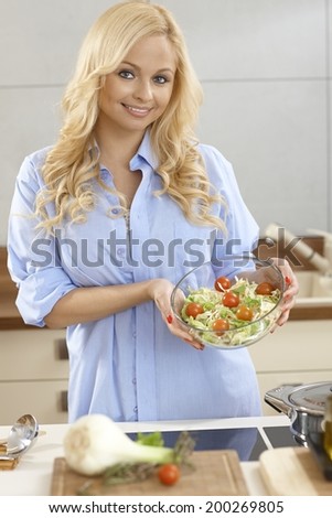 Attractive young blonde woman holding salad bowl, making salad, smiling.