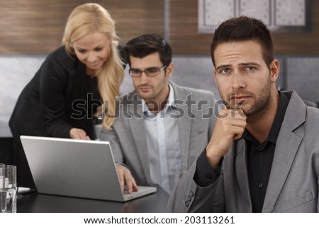Troubled businessman sitting in office, looking troubled, frowning while colleagues working at background.