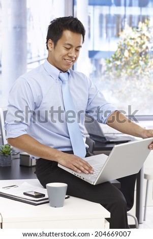 Businessman sitting on top of desk, using laptop computer in bright office.