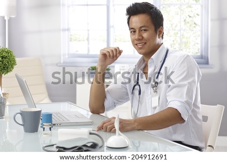 Portrait of happy young doctor sitting at desk, doing paperwork, smiling, looking at camera.