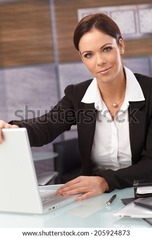 Attractive young businesswoman shutting down laptop, looking at camera, sitting at desk.