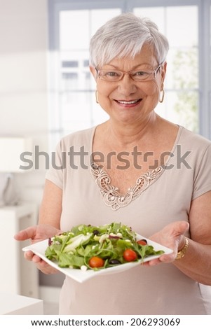 Happy old lady holding plate of fresh green salad, looking at camera.