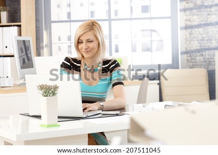 Casual blonde caucasian secretary working with laptop computer in office, smiling.