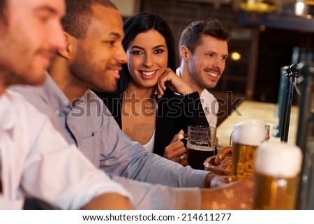 Beautiful young woman sitting at pub with friends, drinking beer. Looking at camera, smiling.