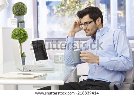 Young businessman sitting at desk, working with laptop computer, drinking coffee, thinking.
