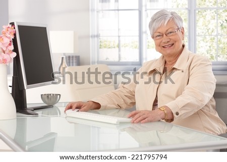 Happy elderly lady sitting at table at home, using computer, smiling at camera.