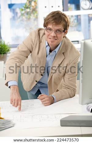 Happy casual engineer pointing at business plan on office table. Leaning on elbow, smiling, looking at camera, wearing glasses, bright.