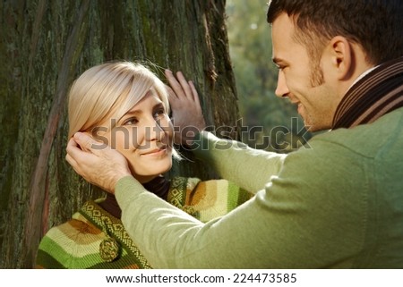Caucasian boyfriend caressing happy romantic blond lady leaning against tree at forest, outdoor. Smiling.