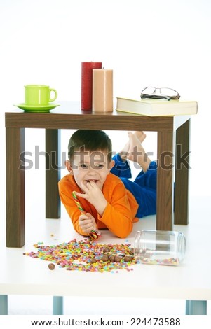Happy little kid eating sweets under table at home. Lying on belly, happy, smiling, isolated on white