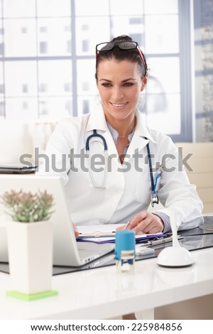Closeup of happy brunette doctor working on laptop computer at medical office, wearing glasses, stethoscope and lab coat, smiling.