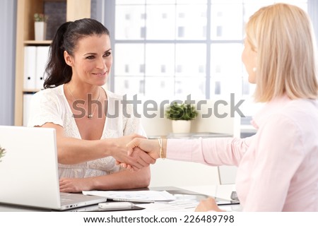 Happy casual brunette businesswoman working with laptop computer, shaking hands with client.