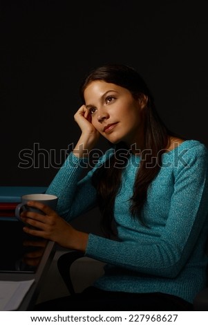 Portrait of young daydreaming woman sitting at desk in dark, looking away, holding tea mug.
