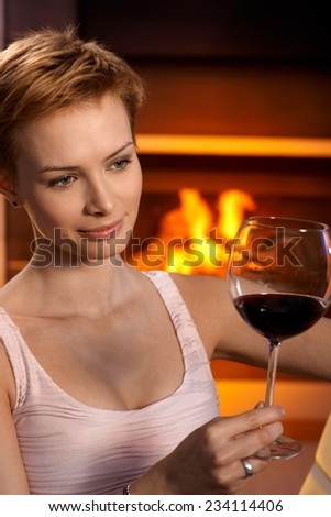 Daydreaming woman looking at glass of wine sitting by fireplace.