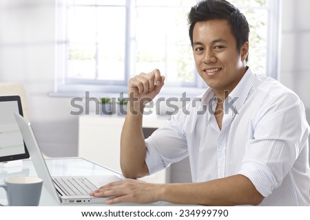 Portrait of happy young Asian businessman sitting at desk, working with laptop computer, looking at camera.