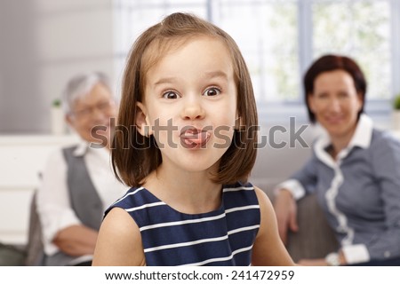 Cute little girl sticking tongue, making funny face, granny and mother looking from background.