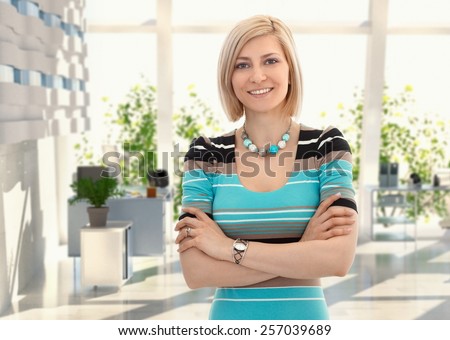 Happy blond female office worker standing arms crossed at office, smiling.
