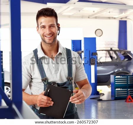 Happy handsome caucasian male car mechanic at auto repair business shop. Wearing workwear clothes and wireless headset, smiling, standing, looking at camera. Greasy, dirty, tablet computer in hand.