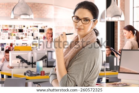 Casual caucasian businesswoman at business startup office with pen in hand, wearing glasses. Looking at camera, scarf around neck.