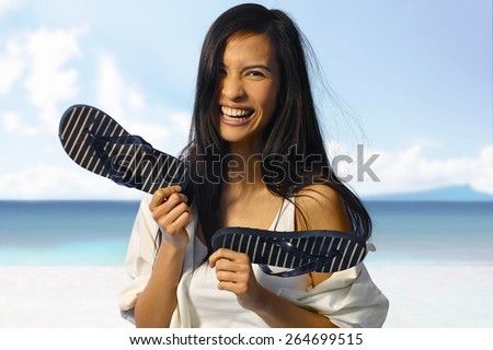 Happy Asian woman laughing on the beach at summertime, holding flip flop in hand.