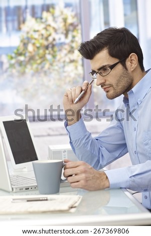 Young businessman sitting at desk, working with laptop computer, thinking.