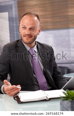 Businessman sitting at desk, holding pen, having personal organizer on desk, smiling, looking away.